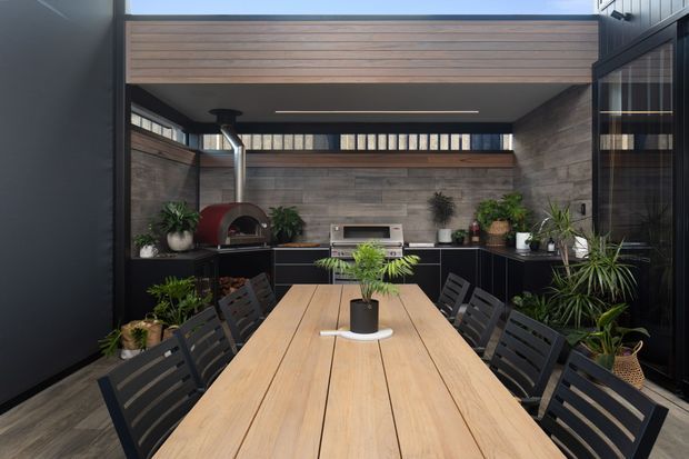 A sophisticated covered outdoor entertaining area with a long timber dining table surrounded by black slatted chairs. The space includes a built-in outdoor kitchen with dark cabinetry, a wood-fired pizza oven, barbecue, timber-lined walls, vertical screening, potted plants, and a large overhead skylight or clerestory opening. The palette is dark and earthy with warm timber accents and lush greenery.