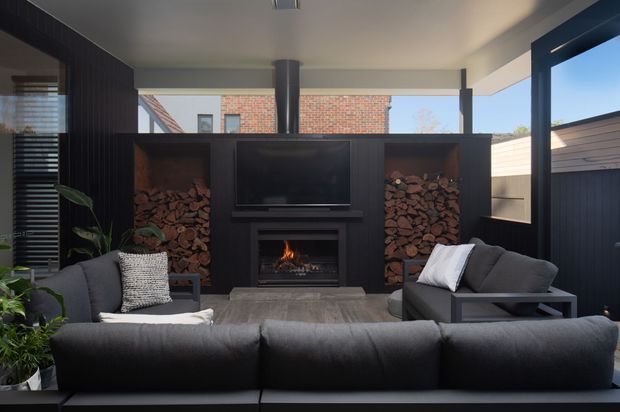 A contemporary indoor-outdoor living room with a large dark sectional sofa facing a built-in entertainment wall. The wall incorporates a television, a linear fireplace, and open timber log storage niches. Full-height glazing and dark-framed openings connect the room to an exterior area, while exposed brick is visible beyond. The palette is predominantly charcoal, black, warm timber, and neutral grey, creating a refined, cosy atmosphere.