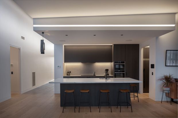 Contemporary open-plan kitchen viewed straight on, featuring a large pale stone island with four dark stools, dark timber cabinetry, integrated appliances, concealed under-cabinet lighting, and light timber flooring. The kitchen opens into a refined living area with a wall-mounted television and indoor plant.