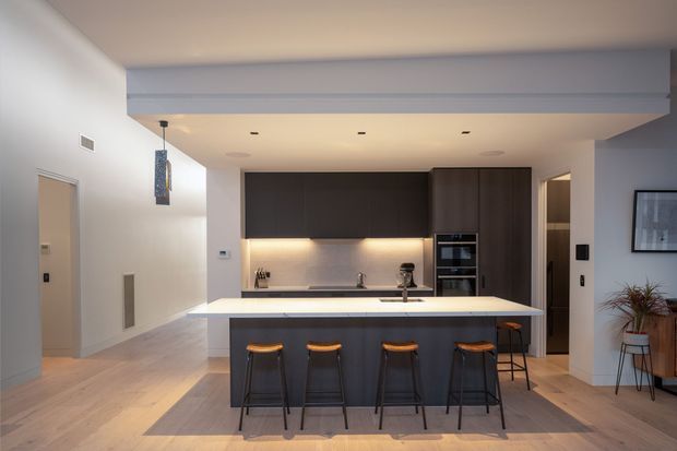 Contemporary open-plan kitchen with a large white island and four dark bar stools, dark wood cabinetry, integrated appliances, under-cabinet lighting, and pale timber flooring. The kitchen opens toward a light-toned living or circulation area, with a framed artwork and plant visible at the side.