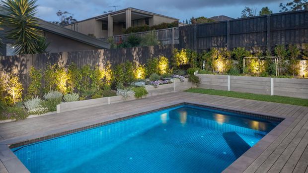 A landscaped backyard with a rectangular swimming pool surrounded by a timber-look deck. Layered garden beds with ornamental plants and shrubs are illuminated by feature lighting, while concrete retaining walls and dark timber fencing define the site. A contemporary house is visible beyond the garden.