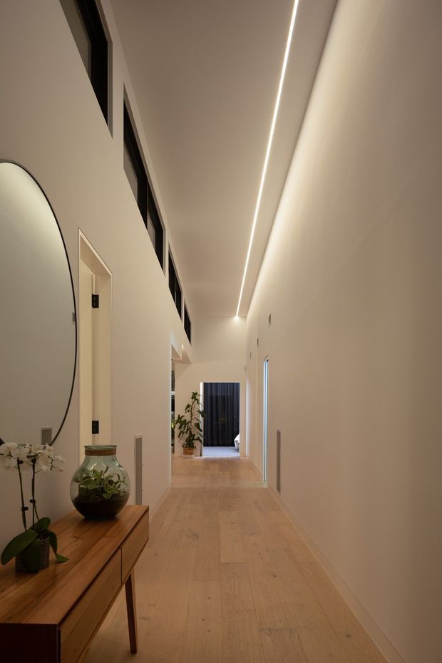 A long, refined residential hallway with pale timber flooring, warm off-white walls, concealed linear ceiling lighting, high-level dark-framed internal windows, and several doorways. A timber console with a round mirror, orchid, and glass planter occupies the foreground, while a plant and dark curtain are visible at the far end.