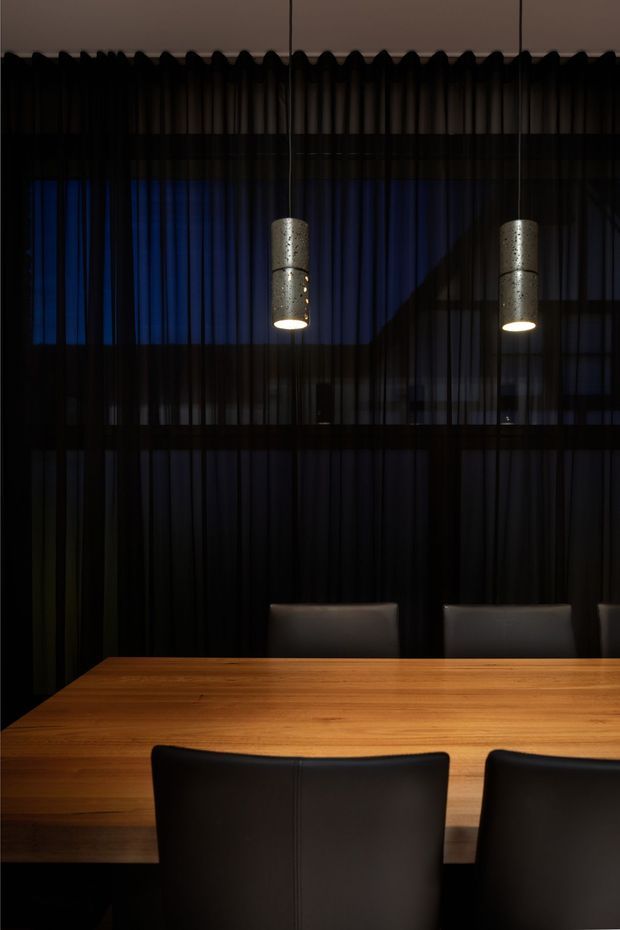 A moody, contemporary dining room centred on a large warm timber dining table with black upholstered chairs. Floor-to-ceiling black sheer curtains create a dramatic backdrop, with deep blue light filtering through glazing and two cylindrical pendant lights providing focused illumination. The space has a refined, intimate atmosphere with dark tones and strong contrast.