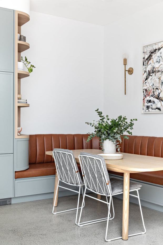 Bright, refined dining nook with a built-in curved banquette upholstered in warm brown leather, integrated muted blue-grey cabinetry and open timber shelving, and a pale timber oval dining table. Two white wire-frame chairs sit on a grey carpeted floor. A brass wall sconce, abstract artwork, and greenery add decorative accents against white walls.