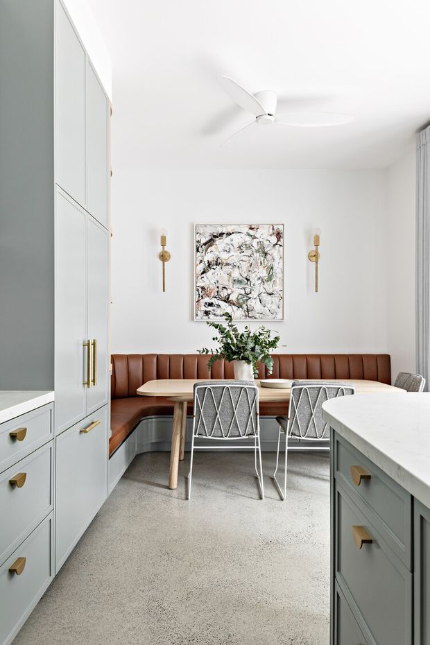 Bright, refined open-plan kitchen dining nook with a built-in cognac leather banquette, round-edged light timber dining table, patterned metal dining chairs, pale blue-grey cabinetry with brass pulls, a white stone island, terrazzo-like flooring, wall sconces, abstract artwork, and a white ceiling fan.