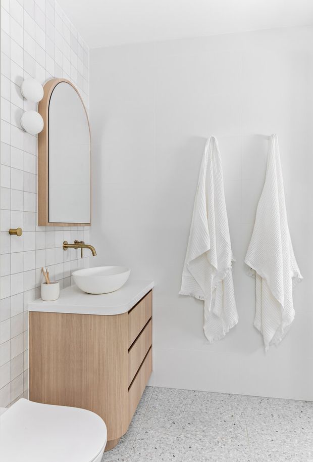 Bright, minimalist bathroom featuring a wall-mounted light timber vanity with white countertop and vessel basin, brushed brass tapware, a tall arched timber-framed mirror, white square wall tiles, white towels, and a pale terrazzo floor. A partial toilet is visible in the foreground.