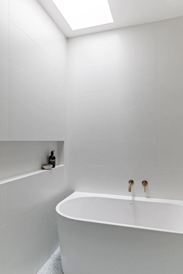 A minimalist contemporary bathroom featuring a freestanding white bathtub, large pale wall tiles, recessed shelving with a small dark bottle, brushed gold wall-mounted tapware, and a large skylight providing soft natural light. A terrazzo-like grey floor is visible at the base.