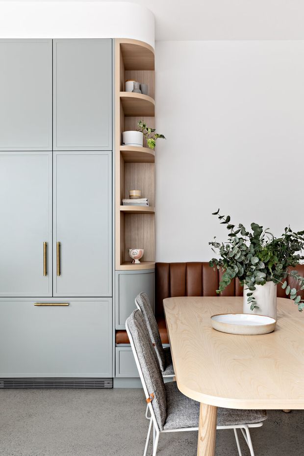 A refined dining area with a built-in pale grey cabinetry wall, integrated storage, and open curved timber shelving. A warm timber dining table with rounded edges is paired with grey upholstered chairs and a brown leather banquette. Brass handles, neutral finishes, and indoor greenery create a calm contemporary interior.