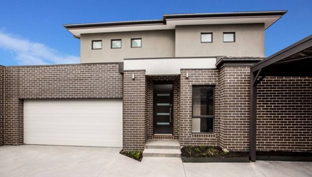 Street-facing view of a contemporary multi-storey townhouse facade with dark brown brickwork, smooth rendered upper walls, a broad low-pitched roof, integrated double garage, covered entry, and a dark glazed front door. The image shows a clean suburban residential exterior under a clear blue sky.