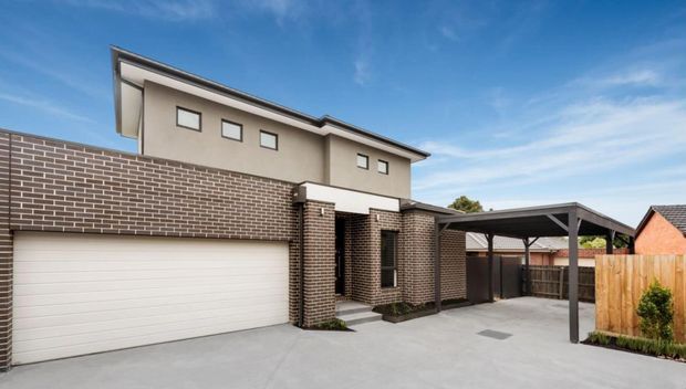 A contemporary multi-storey residential exterior viewed from the driveway, featuring a pale rendered upper level, dark brick lower facade, a wide white sectional garage door, and a covered carport extending to the right. The property has a flat-to-low-pitched roofline, small horizontal windows, concrete paving, and timber fencing under a clear blue sky.