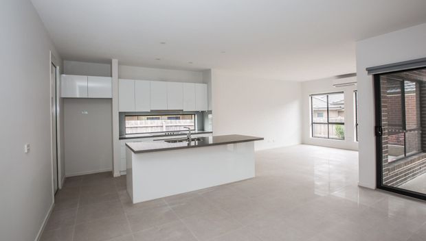 Bright, open-plan contemporary kitchen and living area with white flat-panel cabinetry, a large central island with a grey stone-look benchtop, tiled flooring, and abundant glazing. The space is unfurnished and connects visually to an exterior balcony or terrace through large windows and a sliding glass door.