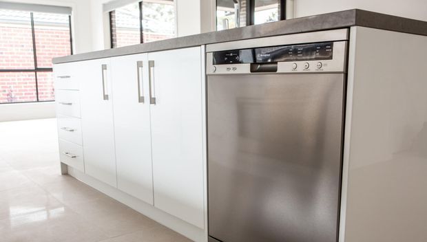 Bright contemporary kitchen interior featuring white flat-panel cabinetry with long brushed-metal handles, a dark grey benchtop, and an integrated stainless-steel dishwasher. Large windows provide abundant natural light, with exposed brick visible outside.