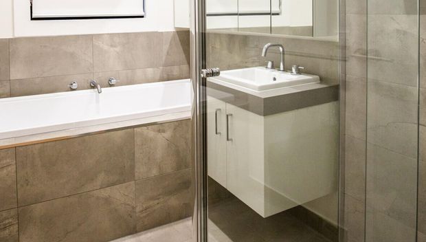 Compact modern bathroom featuring a white bathtub, wall-mounted white vanity with integrated basin, chrome tapware, large beige-grey stone-look wall tiles, and a full-height glass shower enclosure with mirrored reflection.