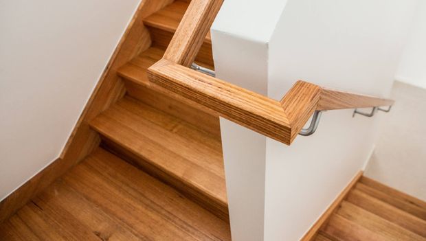 Interior view of a timber staircase turning around a white central wall, with warm wood treads, matching timber handrails, and exposed metal handrail brackets.