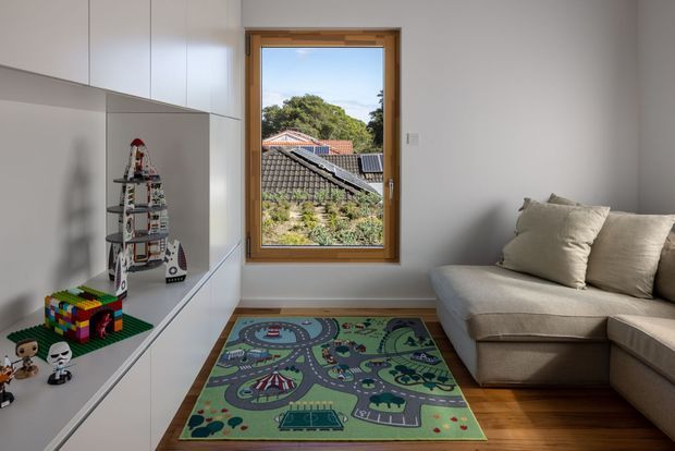 A bright, compact contemporary family room or children's play area with white built-in cabinetry along one wall, a beige sectional sofa, warm timber flooring, and a colourful road-pattern play rug. A timber-framed window provides a view of neighbouring rooftops and greenery. Toys and model structures are displayed on the built-in shelving.