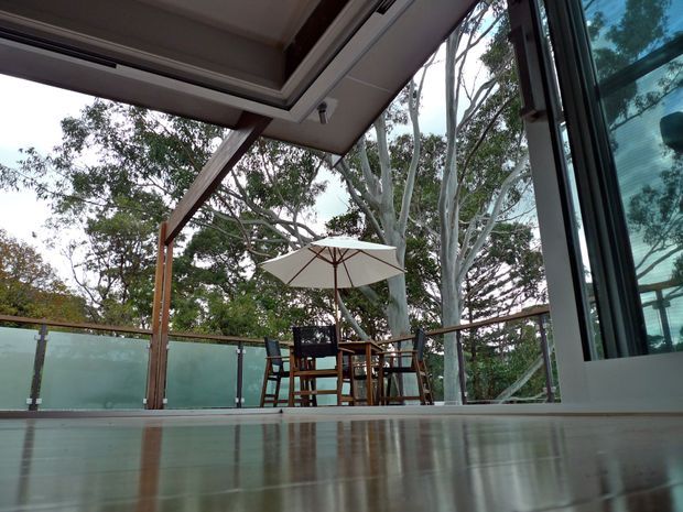Low-angle view from inside a contemporary home toward a covered outdoor dining terrace. The terrace has timber posts and beams, a glass balustrade, timber dining furniture, and a large umbrella, with mature trees visible beyond. Large glazed sliding doors frame the right side, and a reflective polished floor extends through the foreground.