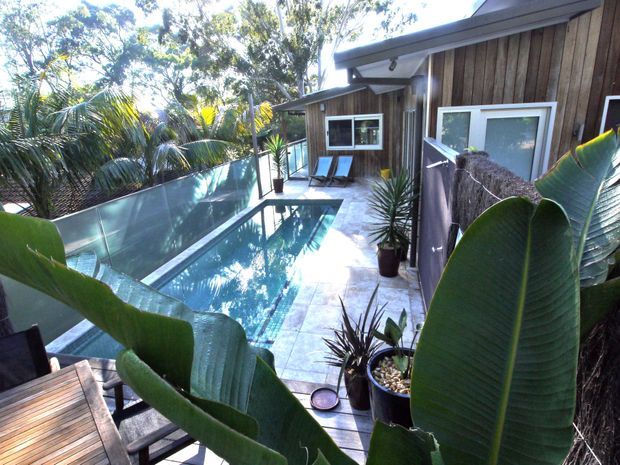 A compact tropical outdoor pool courtyard beside a timber-clad building. The narrow rectangular pool is bordered by pale stone or concrete paving, a translucent privacy screen, timber-framed outdoor seating, and lush banana and palm planting. The building has dark vertical timber cladding, a shallow pitched roof, and simple white-framed windows and doors.