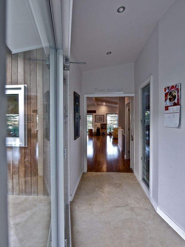 A narrow residential entry hall viewed from beside a large glazed entry door. The hall has pale tiled flooring, white painted walls and ceiling with recessed downlights, a glazed internal door or window on the right, and a timber-clad wall visible through the entry glazing on the left. It opens directly into a warmer living area with polished dark timber flooring and framed artwork.