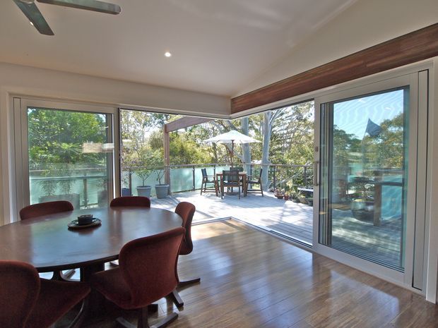 Bright dining area opening onto a spacious timber deck through extensive sliding glass doors. The room features a large dark dining table with upholstered red chairs, warm timber flooring, white walls and ceiling, and a dark timber-toned overhead beam or pelmet. Outdoor dining furniture, an umbrella, glass balustrade and surrounding greenery are visible beyond the glazing.