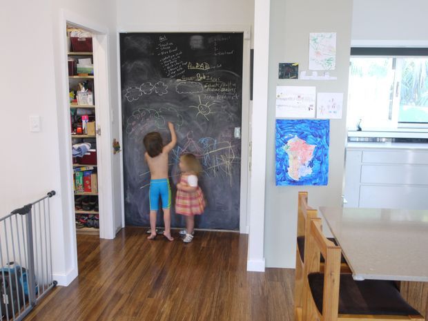 Bright family home interior showing a children's chalkboard wall beside an open pantry or storage cupboard. Two children are drawing on the chalkboard, with artwork displayed on the adjacent wall. The space has timber flooring, pale painted walls, and a partially visible dining area and kitchen with timber chairs and light cabinetry.