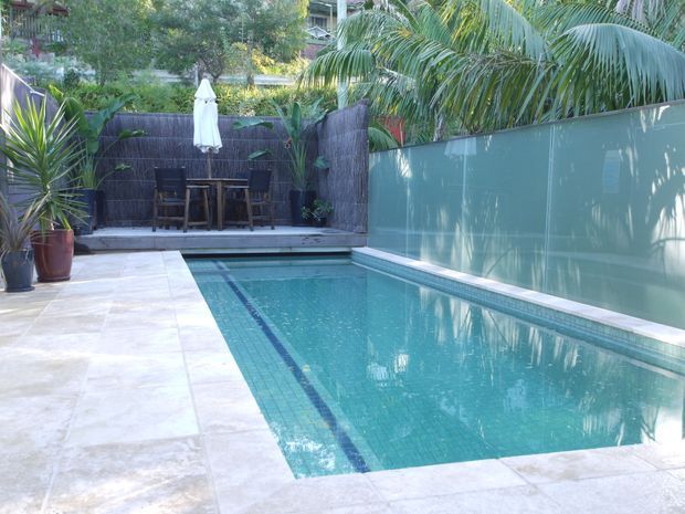A compact outdoor swimming pool courtyard with pale stone paving, turquoise tiled water, and a narrow dark lane detail. A raised timber deck at the far end provides a small dining or lounge area with a table, chairs, and closed umbrella, screened by dark woven fencing and dense tropical planting. A tall translucent privacy fence lines one side, with palm fronds and other greenery creating a secluded resort-like setting.