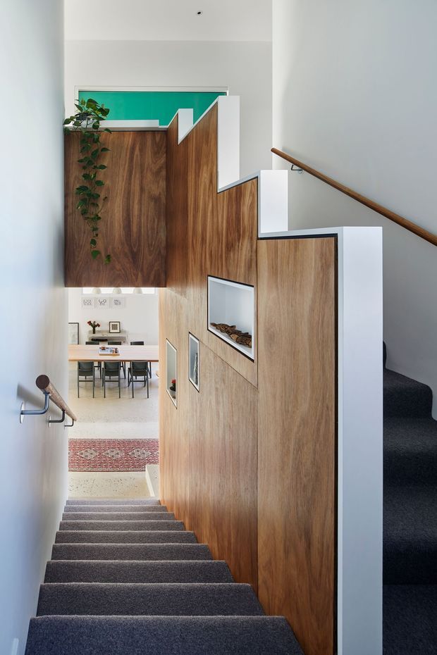 A bright, contemporary stairway with grey carpeted steps, white walls, a timber handrail, and a striking sculptural timber privacy balustrade or partition. The timber joinery incorporates framed display niches and partially screens a dining area below, which contains a light timber dining table, dark chairs, artwork, and indoor plants. A vivid turquoise panel is visible at the upper landing, adding a bold accent.