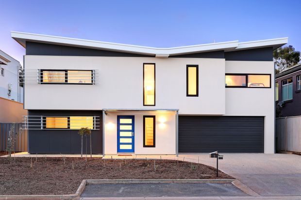 Contemporary two-storey residential facade with crisp white and charcoal cladding, a shallow asymmetrical roofline, horizontal metal balcony rails, narrow vertical windows, a blue entry door, and an integrated double garage. The front garden is minimally landscaped with exposed soil and young trees.
