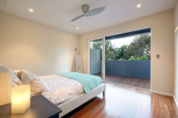 Bright contemporary bedroom with a timber floor, neutral walls, a grey upholstered bed, and a turquoise throw. Full-height sliding glass doors open to a timber deck with a blue-grey privacy screen and leafy greenery beyond. Recessed ceiling lights and a modern ceiling fan complete the room.