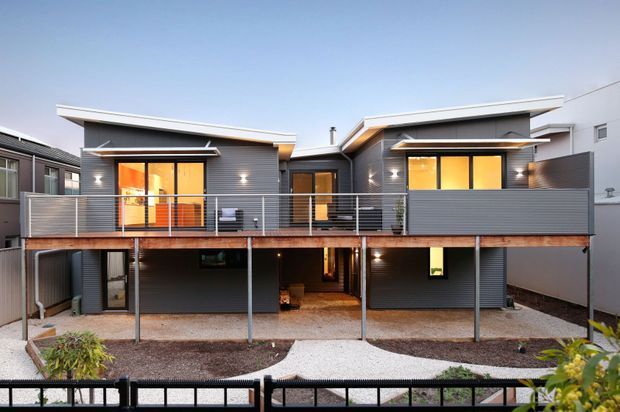 Exterior view of a contemporary multi-level residence with dark horizontal cladding, a broad elevated timber deck, slim metal balustrades, and warm-lit glazing. The split roof forms and projecting upper-level volumes create a modern, articulated facade, with an open undercroft and landscaped frontage below.