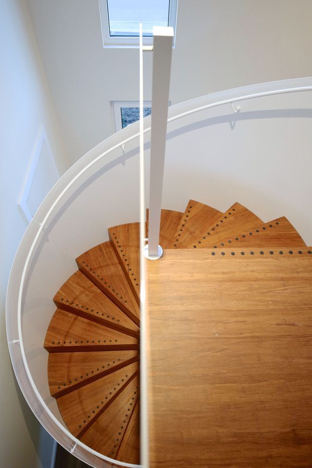Top-down view of a compact spiral staircase with warm timber treads featuring dark circular inlays and a white central newel and curved balustrade. The stairwell has white walls, a skylight, and bright natural light.