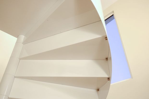 Close-up view looking upward at a white curved spiral stairway with broad wedge-shaped treads, a rounded central support, and small metal tread brackets. A tall skylight or roof window beside the stair provides blue daylight. The image is minimal and architectural, focused on the stair construction and overhead light.