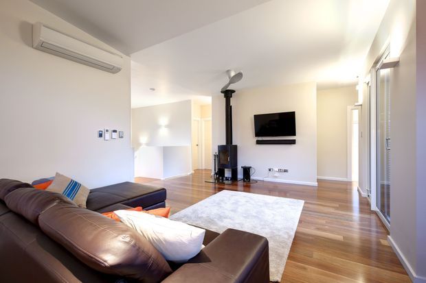 Bright, open-plan contemporary living room with warm timber flooring, white walls and ceiling, a large brown leather sectional sofa, pale area rug, wall-mounted television, and a freestanding black wood-burning stove. Full-height glazed doors or windows line the right side, bringing in abundant natural light. A compact kitchen or utility area is partially visible beyond the living space.