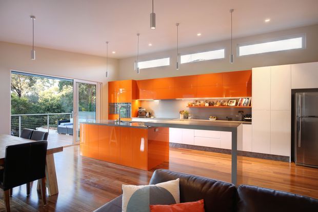 Bright open-plan kitchen and living area with vivid glossy orange built-in cabinetry, a large island, dark stone-look benchtops, warm timber flooring, and integrated stainless-steel appliances. The space has high ceilings with pendant lighting, clerestory windows, and full-height glazing opening to a balcony or deck with leafy views. A dining area and sofa are visible in the foreground.