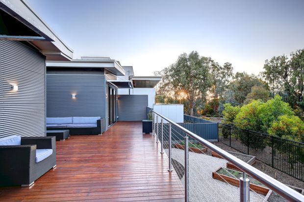 A contemporary elevated outdoor deck or terrace beside a modern multi-level home. The deck has warm reddish timber decking, dark grey horizontal corrugated cladding, outdoor lounge furniture, stainless-steel and mesh balustrades, and integrated raised planters. Native trees and landscaped greenery surround the property beyond the railing.
