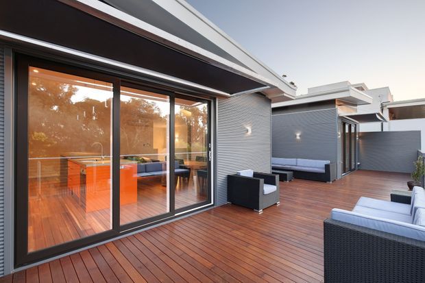 Contemporary outdoor deck and entertaining area adjoining a glazed interior living and dining space. The scene features expansive dark-framed sliding glass doors, warm timber decking, charcoal horizontal cladding, built-in outdoor bench seating, and outdoor lounge furniture beneath projecting roof forms.