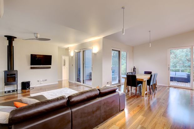 Open-plan contemporary living and dining area with polished timber flooring, a dark leather sectional sofa, freestanding wood-burning stove, wall-mounted television, dining table, and extensive glazed doors and windows opening to an outdoor area. The room has neutral walls, recessed and pendant lighting, and a bright, spacious feel.