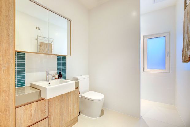 Bright contemporary bathroom with a wall-mounted white basin, timber-look vanity cabinetry, mirrored overhead cabinet, white toilet, pale tiled walls and floor, teal vertical accent tiles around the vanity, and a frosted window. A partial bathtub is visible at left.