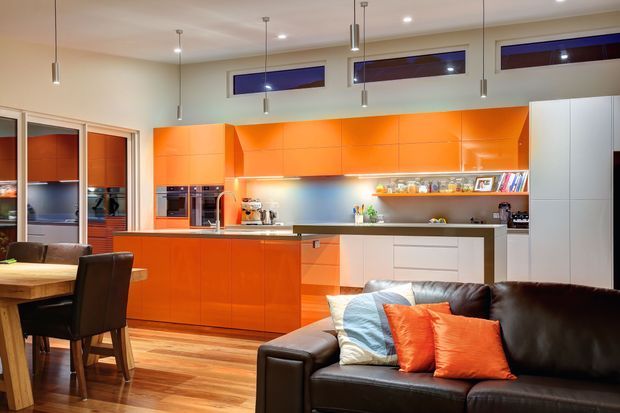 A bright open-plan interior featuring a vivid orange kitchen with extensive flat-panel cabinetry, integrated appliances, a metallic splashback, open display shelving and a central island. A timber dining area and dark leather sofa with orange and blue cushions occupy the foreground, while narrow clerestory windows and slim pendant lights add architectural detail.