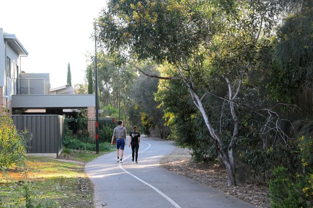 A tree-lined paved shared path curves through a leafy residential area. A contemporary multi-storey home with a grey metal-clad carport, balcony and exterior privacy screens is visible on the left, while dense vegetation borders the path on the right. Two pedestrians are walking along the marked path.