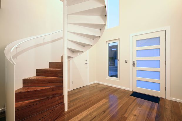 Bright contemporary entry hall featuring a sculptural white spiral staircase with dark timber treads, warm timber flooring, cream walls, a narrow vertical window, and a modern white door with horizontal frosted-glass panels.