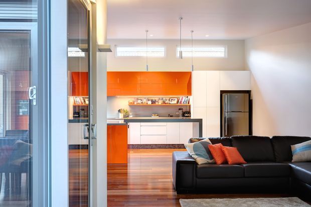 A bright contemporary open-plan living room and kitchen. The foreground features a large black sectional sofa, while the background has a white kitchen with vivid orange glossy cabinetry, a central island, stainless-steel refrigerator, timber flooring, and slim pendant lights. Glazed doors and reflective surfaces appear along the left side.