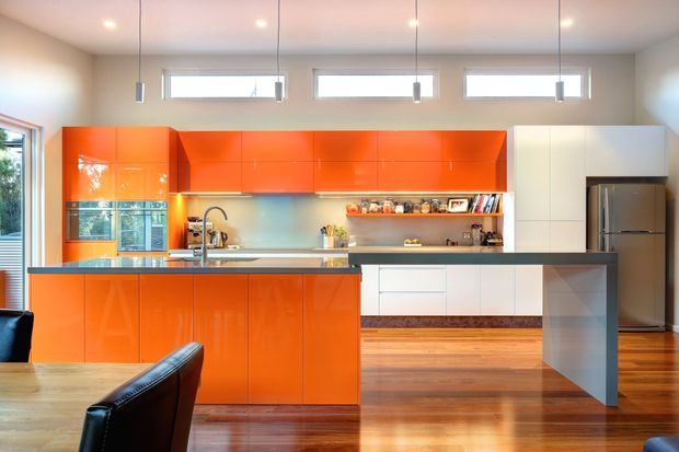 Bright contemporary kitchen with a large glossy orange island and matching upper cabinetry, white built-in cabinetry, stainless-steel refrigerator, open shelving, and a dark benchtop. Warm timber flooring, pendant lights, and high horizontal windows complete the open-plan space.