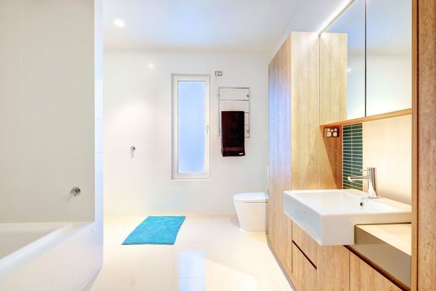Bright contemporary bathroom with a white bathtub at left, wall-hung toilet, compact frosted window, and a floating timber-veneer vanity with an integrated white basin. A narrow green tile splashback and large mirror add colour and reflect the vanity. The space has clean lines, pale finishes, and recessed lighting.