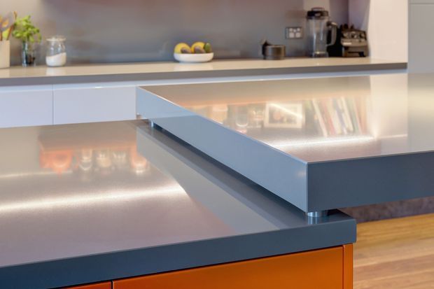 Close-up of a contemporary kitchen island and benchtop arrangement, featuring layered glossy grey surfaces, dark blue-grey edging, integrated cabinetry, warm under-bench lighting, and bright orange cabinet fronts. A second kitchen counter with small appliances and open shelving is visible in the background.