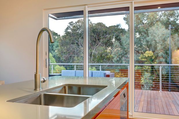 Bright contemporary kitchen interior focused on a large stainless-steel double-bowl sink and curved faucet set into a pale stone island benchtop. Full-height glazed sliding doors open to a timber deck with slim metal balustrading and leafy vegetation beyond. Glossy orange cabinetry adds a strong accent beneath the island.