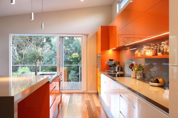 A bright contemporary kitchen with glossy orange-red cabinetry, white lower cabinets, pale stone benchtops and timber flooring. The room features a central island with an integrated sink and curved tap, pendant lighting, open shelving with kitchenware, and full-height cabinetry along one wall. Glazed doors open to a timber balcony or deck overlooking trees.