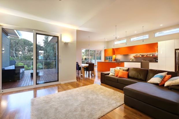 Open-plan contemporary living area with a dark brown sectional sofa, large neutral rug, timber flooring, and a striking orange kitchen with integrated white cabinetry. Full-height glazed doors open to a timber deck and greenery, while a dining table sits between the living and kitchen zones. High-level windows and recessed lighting create a bright, connected interior.