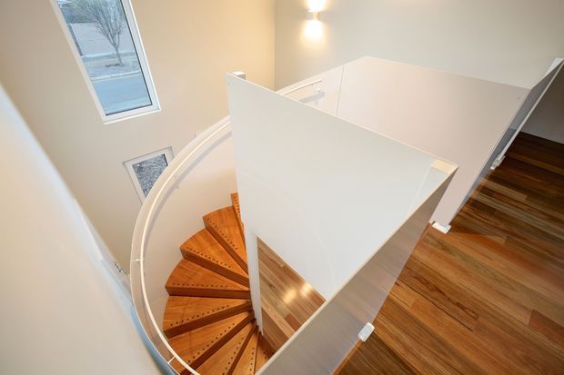 High-angle interior view of a modern curved staircase with warm timber treads and a white balustrade, opening onto a polished timber-floored landing. Tall windows bring natural light into the stairwell.