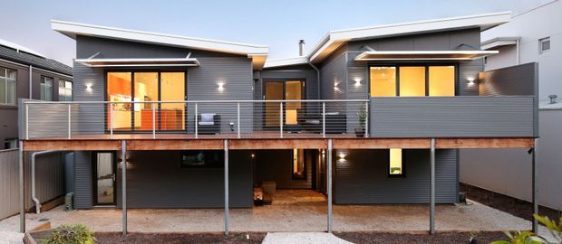Exterior view of a contemporary multi-level residential dwelling with dark horizontal cladding, asymmetrical skillion-style roof forms, warm-lit windows, a long upper-level balcony with metal balustrade, and an open ground-level area beneath the elevated living spaces.