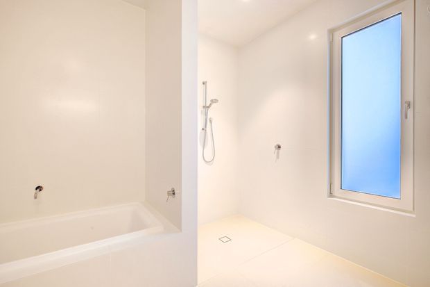 Bright, minimal white bathroom showing a bathtub on the left and an open walk-in shower area with handheld shower fittings, floor drain, and frosted window. The space has clean tiled surfaces and a simple contemporary fit-out.